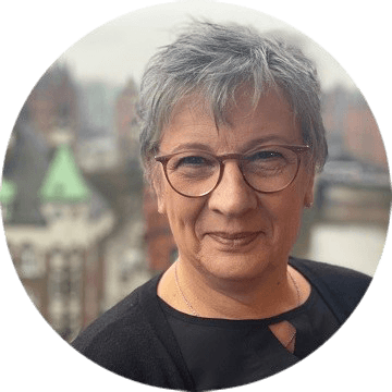 Porträt einer Frau mit kurzen grauen Haaren und Brille vor urbaner Kulisse