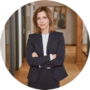 Frau mit schulterlangem Haar im dunkelblauen Anzug steht mit verschränkten Armen im Büro