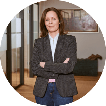 Frau mit mittellangem braunem Haar und grauem Blazer steht im Büro mit verschränkten Armen