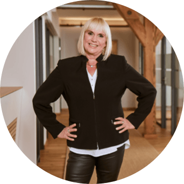 Blondhaarige Frau im schwarzen Blazer posiert im Büro mit den Händen in der Hüfte