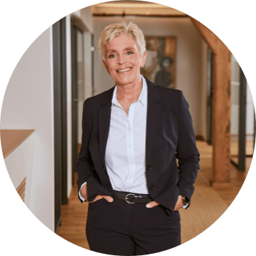 Frau mit kurzem, hellblondem Haar und dunkelblauem Hosenanzug im modernen Büro