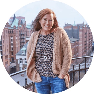 Frau mit langem, braunem Haar und beigefarbener Strickjacke auf Balkon mit Blick auf die Speicherstadt