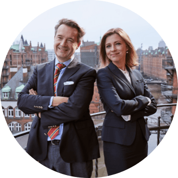 Zwei Geschäftsleute im Anzug mit verschränkten Armen auf einer Terrasse mit Speicherstadt im Hintergrund