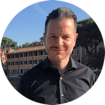 Mann mit dunklem Hemd vor mediterranem Gebäude und blühenden Bäumen