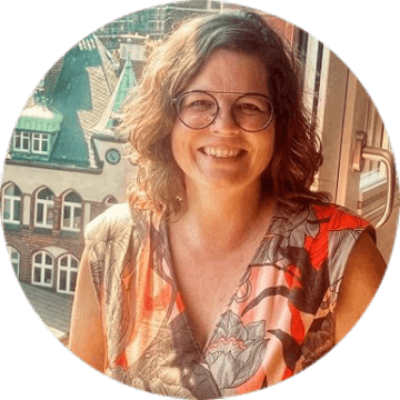 Frau mit lockigem Haar und Brille vor Fenster mit Blick auf historische Gebäude