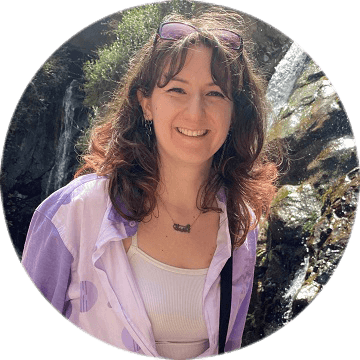 Frau mit dunklen Locken in lilafarbener Bluse steht lächelnd vor einem Wasserfall