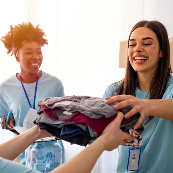 Zwei junge Frauen in blauen T-Shirts nehmen lachend Kleiderspenden entgegen