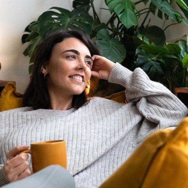 Frau mit dunklem Haar und Kuschelpullover entspannt mit einer Tasse Tee auf dem Sofa